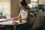 A single parent sitting at a sunny California kitchen table reviewing bills and finances, with a child’s art on the wall and light streaming through an open window, symbolizing hope and support.