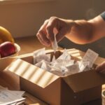 A parent sorting household receipts into a small box on a kitchen counter, surrounded by everyday items like fruit, school forms, and coffee, with sunlight streaming through blinds — a real-life moment that reflects practical, zero-fee budgeting and the simplicity of Mint alternatives that cost nothing.