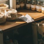 A realistic kitchen counter with open bags of rice, mason jars, and a handwritten grocery list, showing a single parent’s practical approach to buying household staples in bulk without a membership fee.